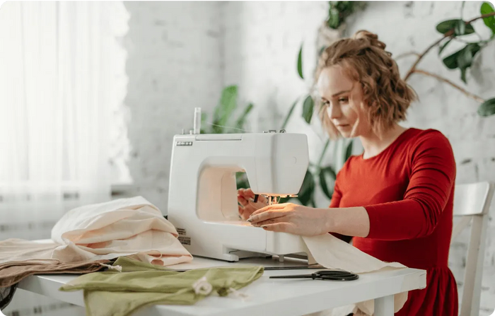 Woman in a red dress using a sewing machine in a bright room with plants.