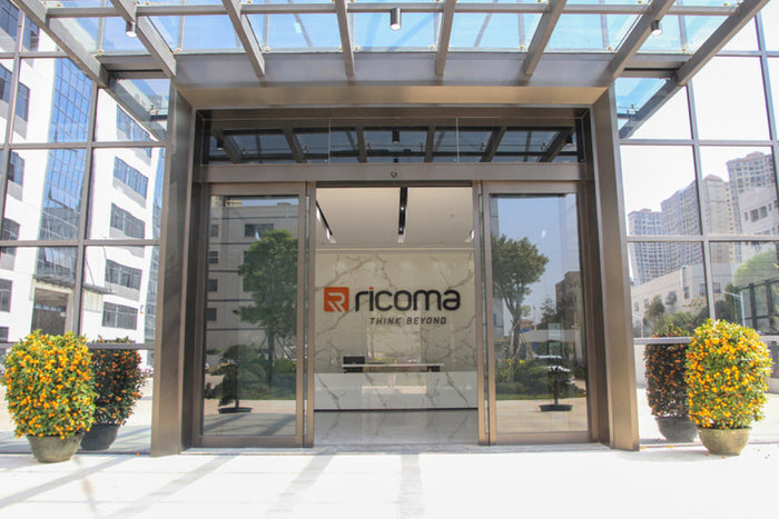 Entrance to a building with Ricoma logo, surrounded by decorative plants.